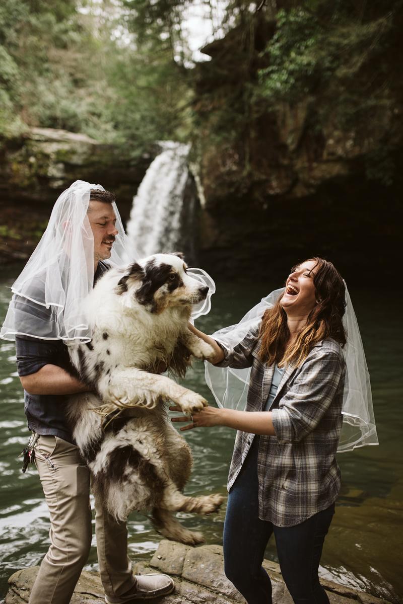 Veronica and Joe hold up their dog in front of a waterfall